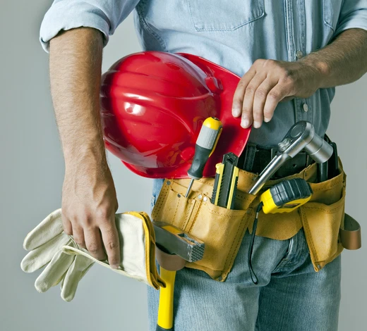 Ouvrier en bâtiment portant casque de sécurité rouge, ceinture porte-outils en cuir avec compartiments multiples, gants de protection beiges. Tient marteau à manche en bois dans une main. Équipement professionnel de construction visible: tournevis, outils de mesure, accessoires de chantier dans la ceinture utilitaire.