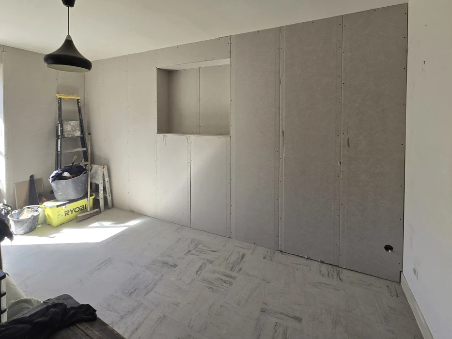 Intérieur en cours de rénovation avec cloisons sèches installées. Échelle métallique, seau de peinture, outils Ryobi au sol. Sol carrelage imitation marbre blanc-gris. Murs en plaques de plâtre BA13 vissées, joints apparents non enduits. Niche murale intégrée. Plafond suspendu avec spots encastrés. Chantier second œuvre plaquiste.