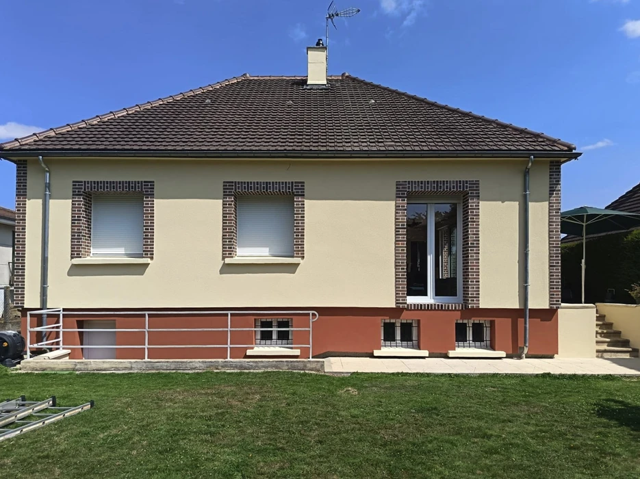Façade d'une maison individuelle contemporaine en cours de finition avec échafaudage. Le niveau supérieur présente un enduit beige avec encadrements en briques et volets roulants blancs, porte-fenêtre vitrée. Le soubassement arbore un enduit rouge orangé avec petites fenêtres à barreaux. Toiture en tuiles mécaniques, gouttières PVC, terrain enherbé en premier plan.