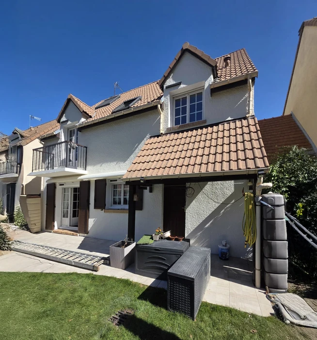 Maison individuelle moderne à étage avec toiture en tuiles terre cuite et façade enduite gris clair. Jardin paysager avec pelouse entretenue, terrasse carrelée et mobilier d'extérieur contemporain.