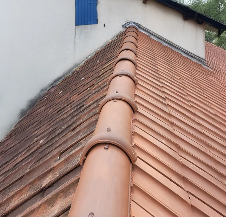 Toiture en tuiles canal terre cuite avec faîtage arrondi, vue en plongée. Couverture traditionnelle méditerranéenne avec tuiles emboîtées et fixations visibles. Faîtage courbe avec tuiles faîtières scellées. Surface uniforme orangée caractéristique de l'argile cuite. Étanchéité assurée par recouvrement des éléments céramiques.