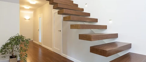Escalier flottant moderne en bois massif avec marches en porte-à-faux fixées au mur blanc. Intérieur contemporain avec parquet assorti, plante verte décorative, portes blanches et éclairage encastré au plafond.