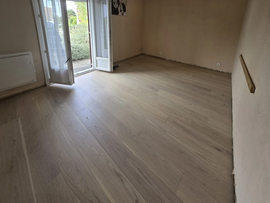 Pièce vide en cours de rénovation avec parquet stratifié beige nouvellement posé. Murs blancs, plinthes en cours d'installation, porte-fenêtre ouverte donnant sur extérieur verdoyant.