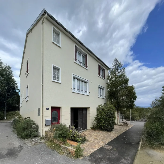 Façade de maison individuelle contemporaine à deux étages avec enduit beige clair. Volets rouges aux fenêtres, porte d'entrée rouge et porte de garage grise au rez-de-chaussée. Environnement résidentiel avec végétation et ciel nuageux.