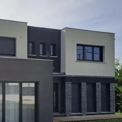 Façade contemporaine de maison individuelle moderne avec architecture géométrique bi-ton. Murs enduit beige et gris anthracite, baies vitrées rectangulaires à châssis noir, volets roulants aluminium. Rez-de-chaussée avec parement pierre naturelle grise, colonnes structurelles, toiture terrasse étanchée membrane EPDM.