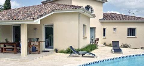 Maison contemporaine avec piscine et terrasse aménagée. Bâtiment de plain-pied en crépi beige, toiture tuiles terre cuite, baies vitrées coulissantes. Espace détente avec transats, mobilier extérieur bois. Margelles piscine carrelage, liner bleu. Aménagement paysager méditerranéen.
