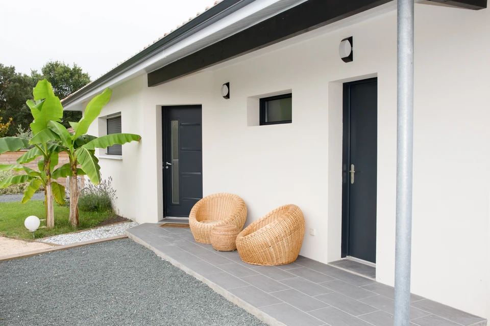 Entrée moderne d'une maison contemporaine avec terrasse couverte. Deux fauteuils en rotin tressé disposés devant la façade. Porte d'entrée anthracite avec insert vitré, fenêtre assortie. Murs enduit blanc, toiture plate débordante. Carrelage gris au sol, colonne structurelle métallique. Aménagement paysager avec bananier et gazon.