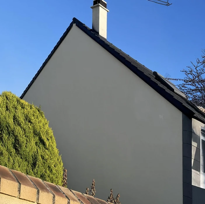 Façade latérale d'une maison résidentielle avec mur pignon blanchi, toiture en tuiles grises et gouttière noire. Environnement extérieur avec végétation de conifères et ciel bleu dégagé.