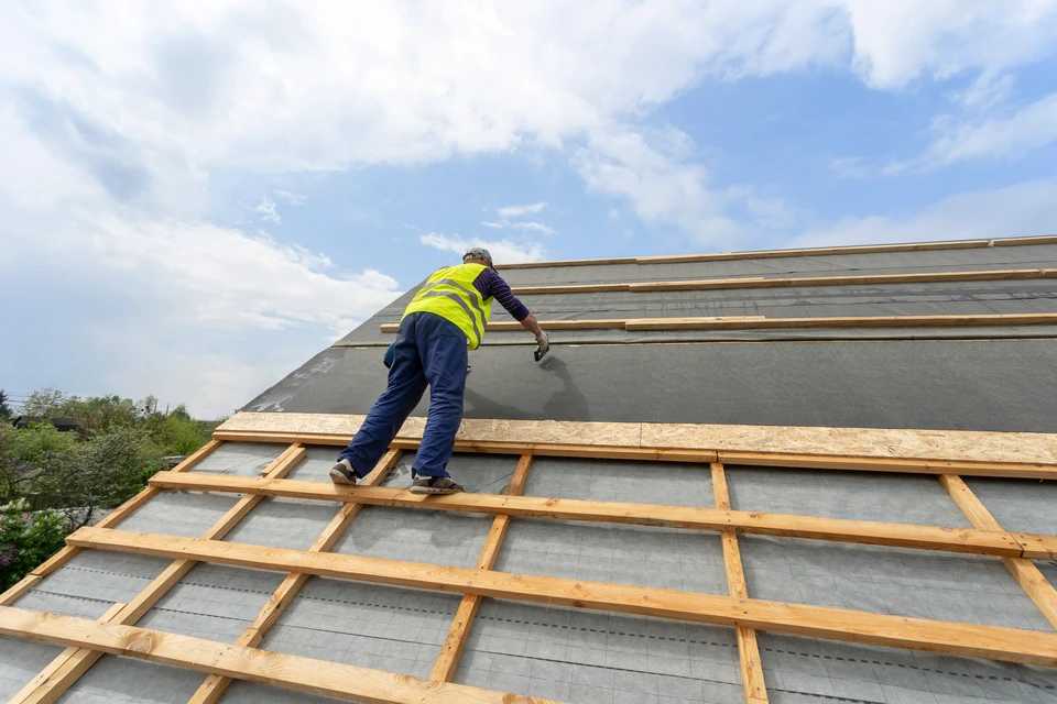 Ouvrier en gilet haute visibilité jaune installant une membrane d'étanchéité sur toiture en pente. Charpente en bois avec liteaux horizontaux, sous-toiture grise respirante. Ciel nuageux, environnement extérieur résidentiel.