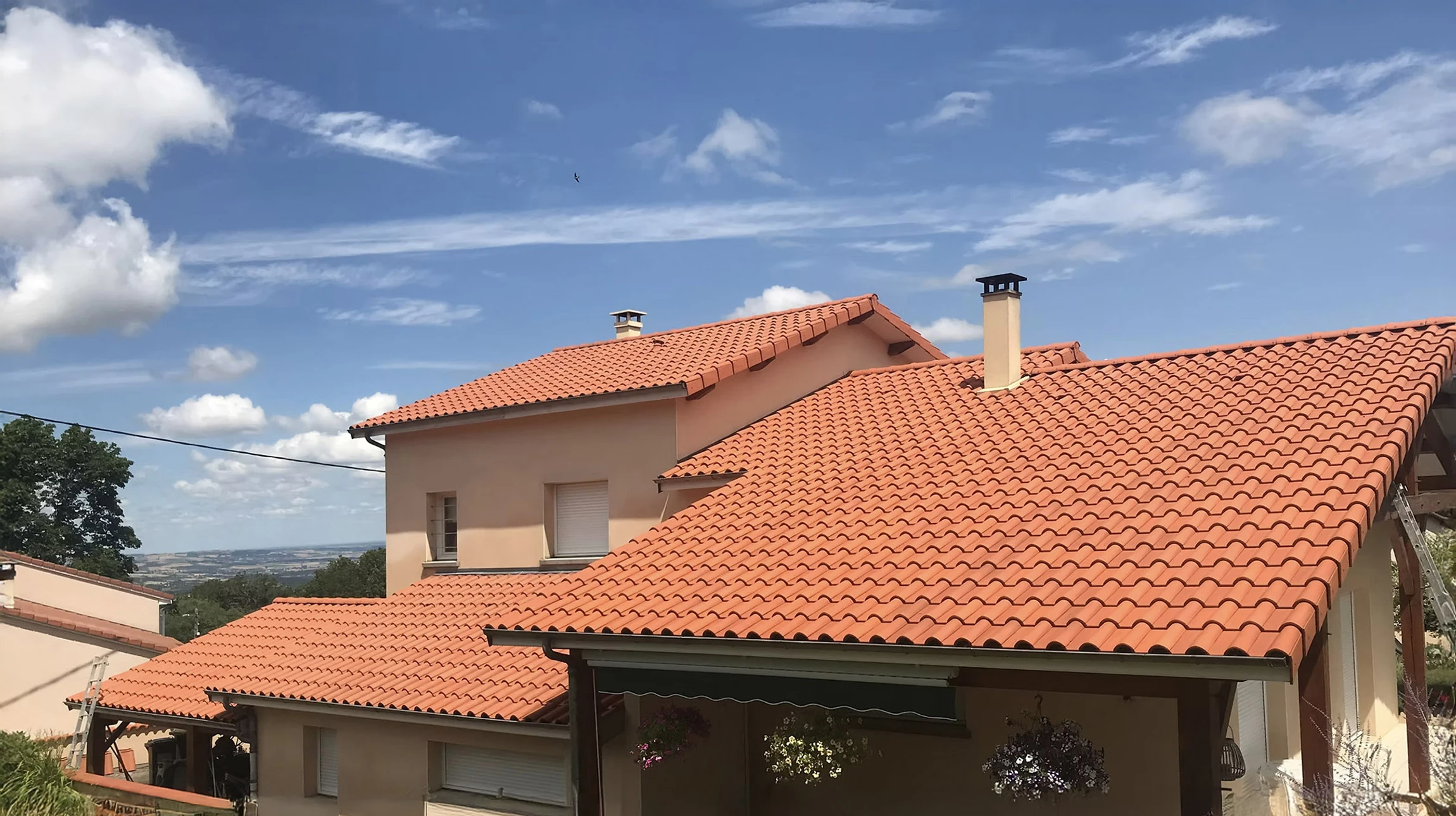 Façade extérieure d'une maison moderne avec toiture en tuiles canal terre cuite et gouttière métallique apparente. Mur crépi beige clair avec encadrement de fenêtre blanc visible partiellement. Ciel bleu dégagé en arrière-plan.