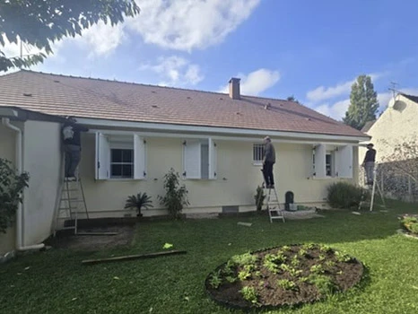 Maison résidentielle avec toiture en tuiles canal terre cuite de couleur rouge orangé sous ciel bleu nuageux. Vue extérieure d'une habitation à étages avec murs enduits beige, volets roulants blancs, cheminées avec mitrons. Couverture en tuiles mécaniques à emboîtement, faîtage traditionnel, descentes pluviales. Architecture méditerranéenne typique.