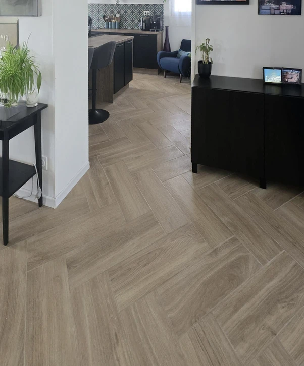 Scène extérieure de pose de carrelage sur terrasse. Un ouvrier utilise une règle de maçon beige pour ajuster l'alignement des dalles grises. Surface partiellement carrelée avec joints réguliers.