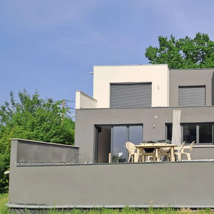 Extérieur d'une maison résidentielle avec lucarne en saillie. Fenêtre à double vitrage avec encadrement blanc et volets blancs à lamelles horizontales. Bardage en bois teinté rouge-brun à lames horizontales avec joints creux. Toiture en tuiles terre cuite de type canal ou romanes, gouttière blanche en PVC. Conifère en premier plan.