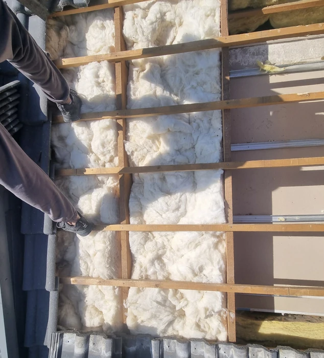 Toiture résidentielle avec tuiles canal en terre cuite rouge-orange disposées en rangées régulières. Fenêtre de toit rectangulaire intégrée avec cadre sombre et verre translucide. Gouttière métallique grise avec supports de fixation visibles le long du débord de toit.