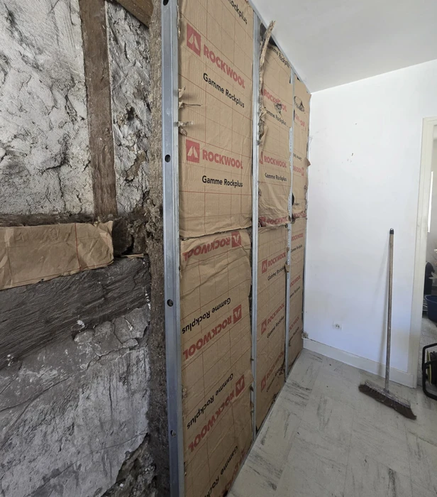 Chantier de construction intérieur avec ouvrier installant l'isolation thermique entre solives de plancher. Travailleur en vêtements de protection manipulant matériau isolant blanc.